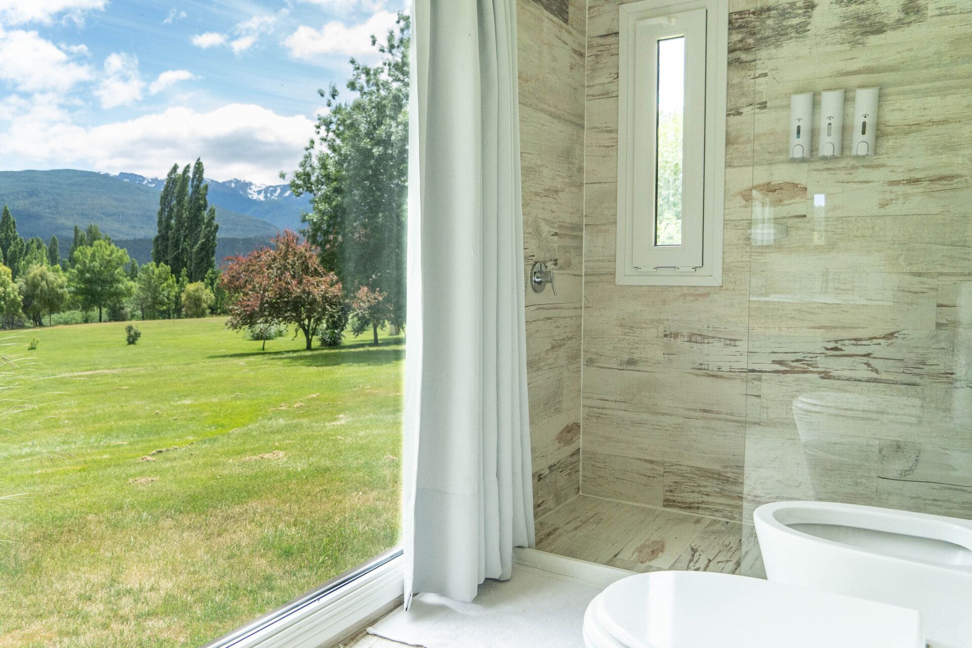 Baño moderno en La Aguada Suite con ducha, revestimiento claro y ventana con vista a montañas en El Bolsón Baño moderno en La Aguada Suite con ducha, revestimiento claro y ventana con vista a montañas en El Bolsón